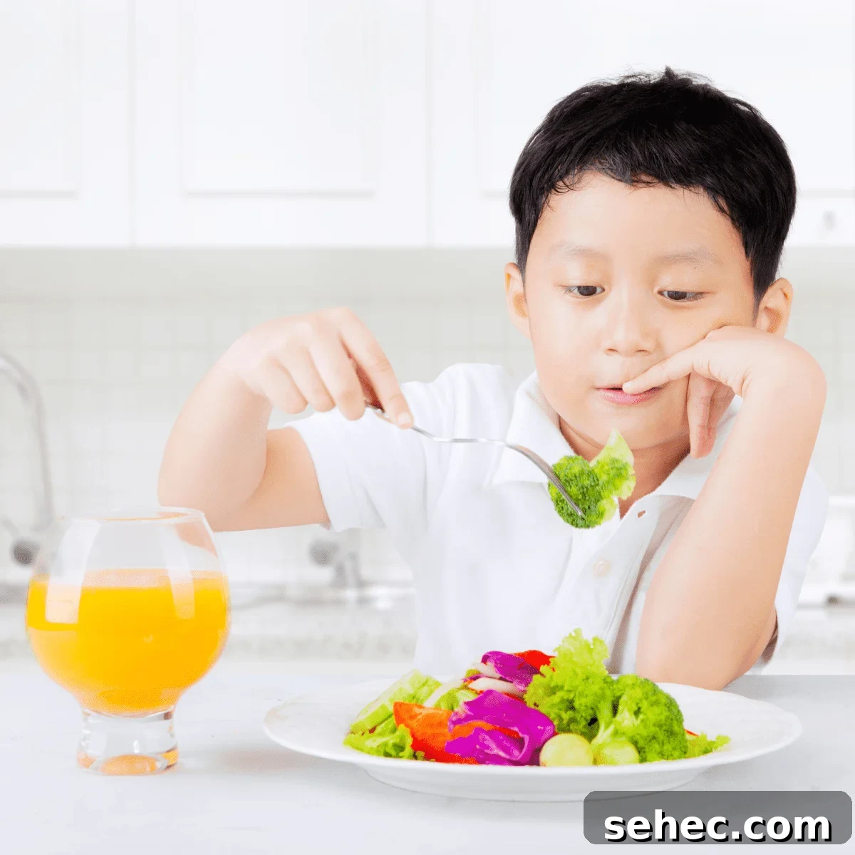 A boy in a white kitchen eating a colorful plate of vegetables with a clear glass of orange juice.