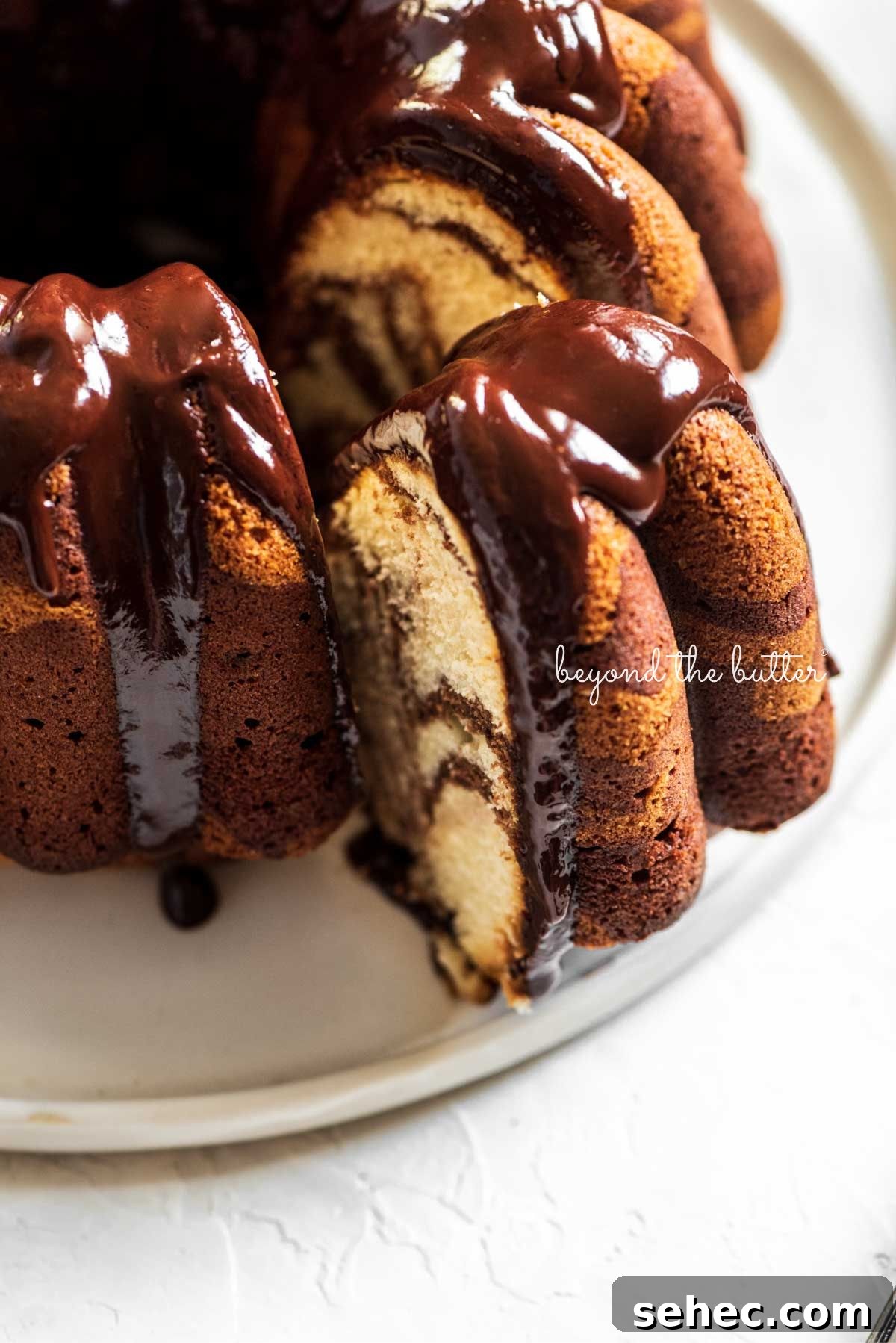 Moist Marble Bundt Cake Recipe 2 Chocolate glazed marble bundt cake with one slice partially removed on a white ceramic dessert plate and white background.