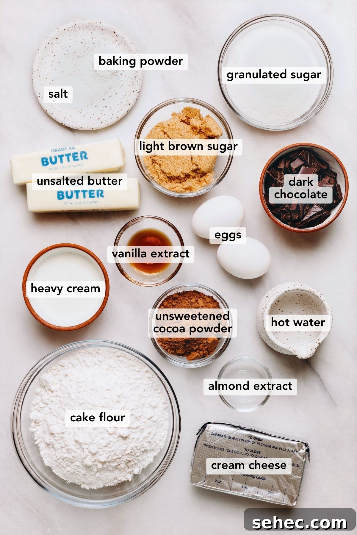Moist Marble Bundt Cake Recipe 4 Ingredients in various size bowls and plates labeled as salt, baking powder, granulated sugar, light brown sugar, unsalted butter, heavy cream, vanilla extract, almond extract, cream cheese, hot water, unsweetened cocoa powder, cake flour, dark chocolate, and eggs on a white marbled background.