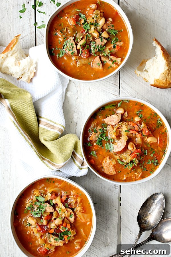Farmhouse Kielbasa Cabbage Soup with Cannellini Beans 5 Photo of three servings of Farmhouse Cabbage Soup with Cannellini Beans and Kielbasa on white wooden background with green towel and broken bread.