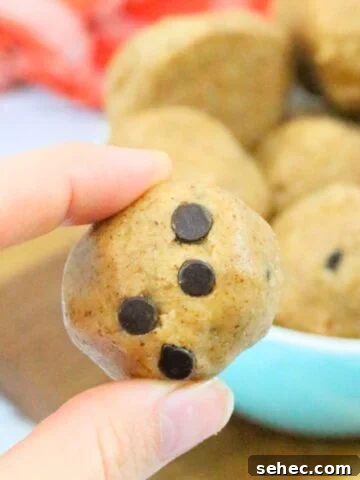 Raw cookie dough ball with chocolate chips being held between two fingers and more balls in a bowl behind them.