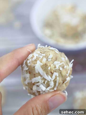 Two fingers holding a coconut protein ball with a purple bowl of almonds and coconut behind it