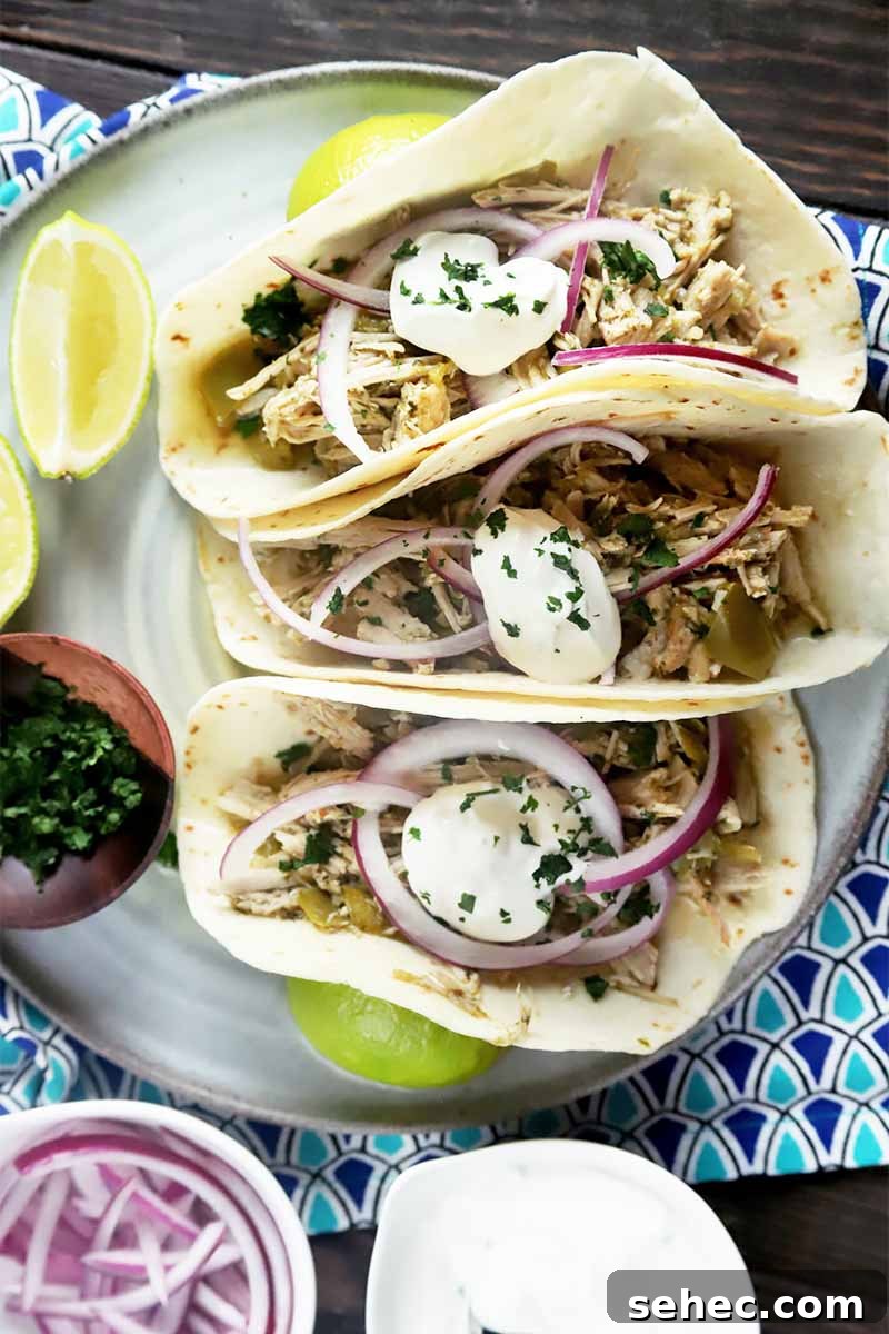 Slow Cooker Pork Tacos for Taco Tuesday 3 Vertical image of three flour tortillas filled with meat, sour cream, and onions on a plate next to limes and a bowl of sliced onions on a blue napkin.