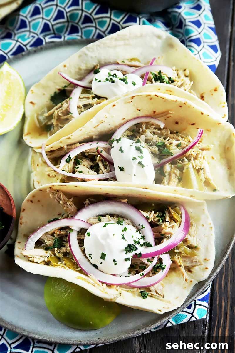 Slow Cooker Pork Tacos for Taco Tuesday 4 Vertical top-down image of three flour tortillas filled with shredded meat, sour cream, sliced onions, and chopped herbs on a plate on a blue patterned napkin.