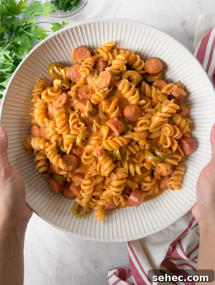 Creamy Hot Dog Pasta Recipe with Tomato Sauce 3 a hand holds a white bowl of creamy hot dog pasta from above
