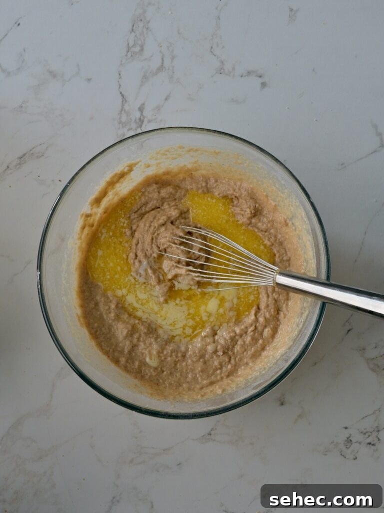 Moist Applesauce Crumb Cake with Cinnamon Streusel 12 Mixing melted butter into the applesauce cake batter with a whisk in a medium mixing bowl on a marble countertop.