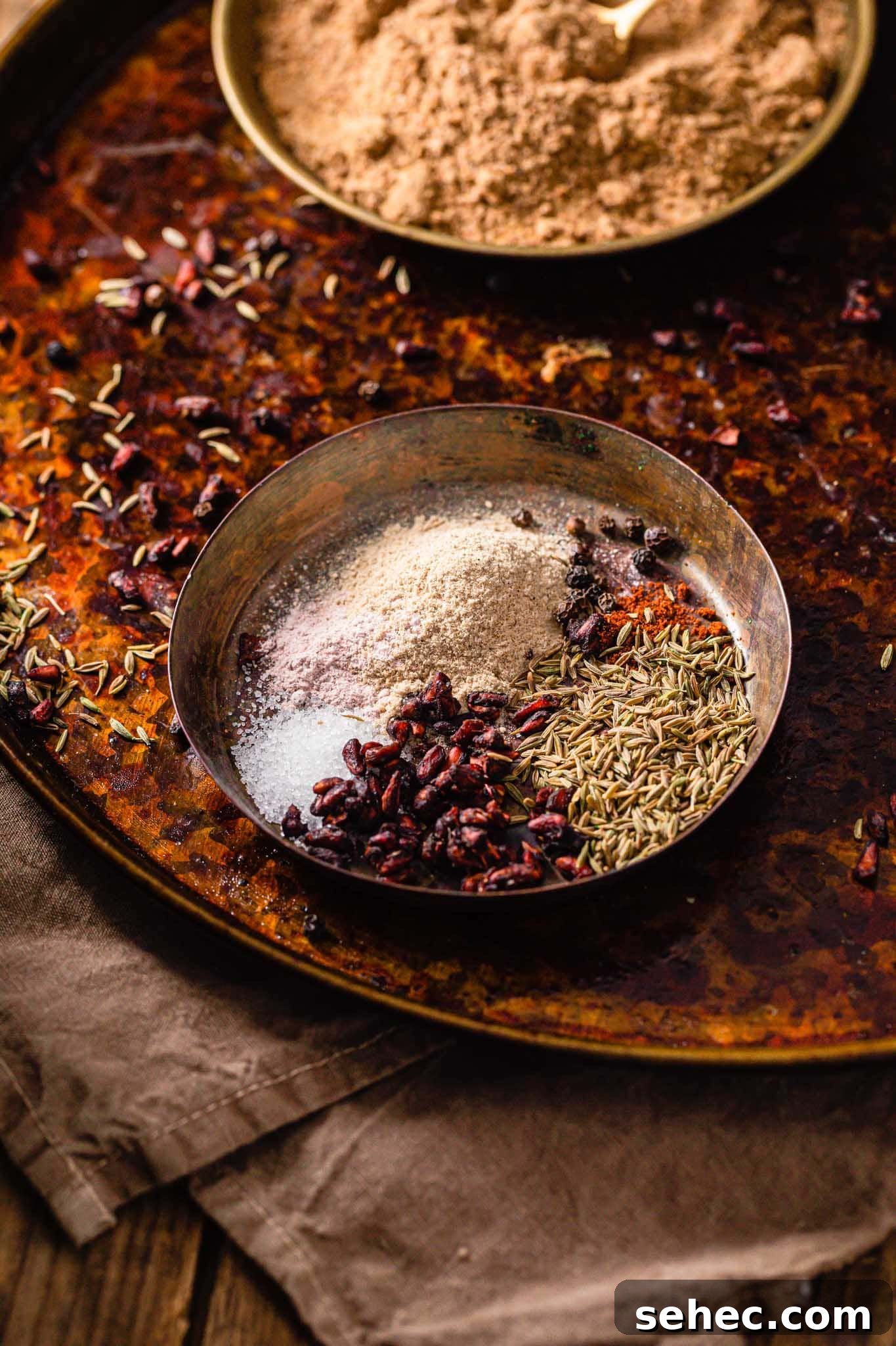 Authentic Chaat Masala Recipe: Homemade Indian Spice Mix 5 A brass bowl containing dried mango powder (amchur), dried pomegranate seeds (anardana), cumin seeds, black peppercorns, red chili powder, and sugar