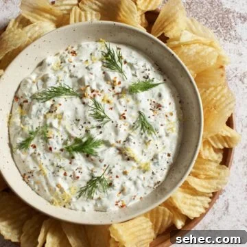 A bowl of dill pickle dip topped with fresh dill, pepper, and lemon zest, surrounded by ridged potato chips.