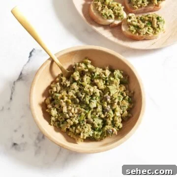 Green olive tapenade in a wooden bowl with a gold spoon in it, a plate of bread with tapenade on it is in the upper right.