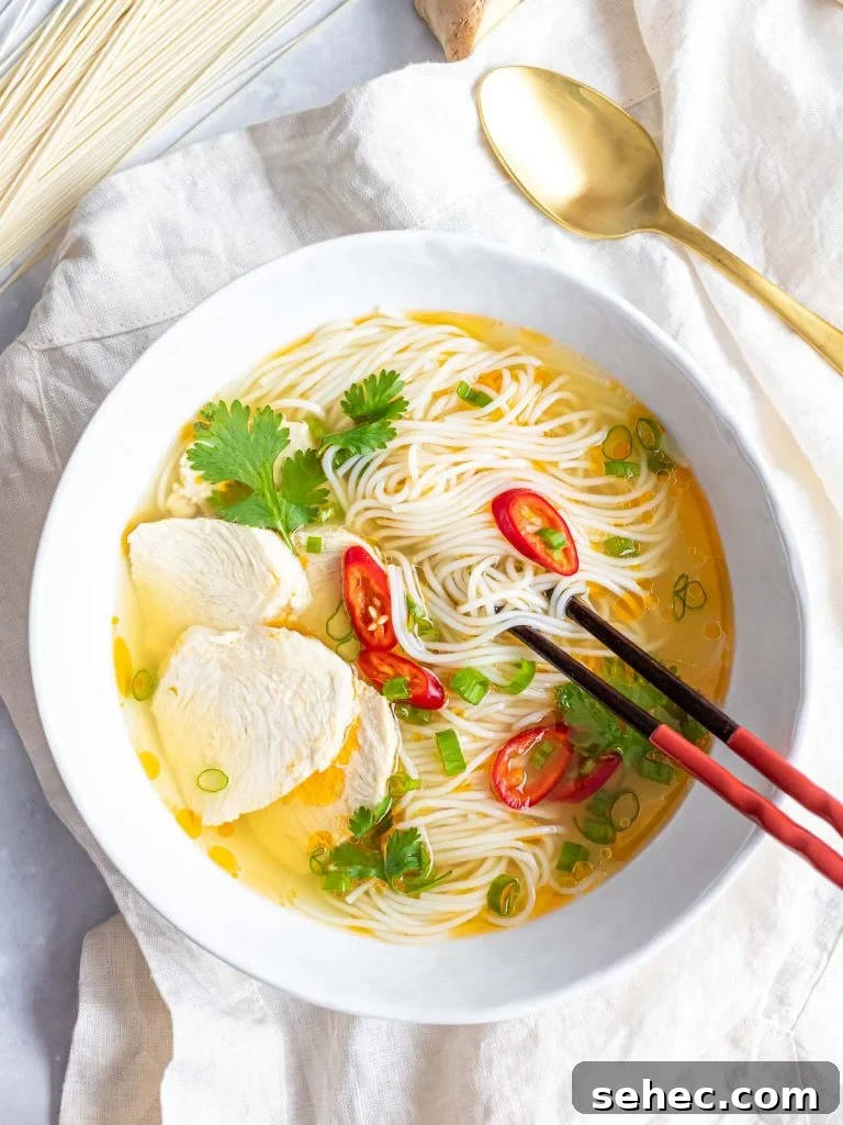 Asian chicken noodle soup with red peppers and herbs in a white bowl with chopsticks and a spoon