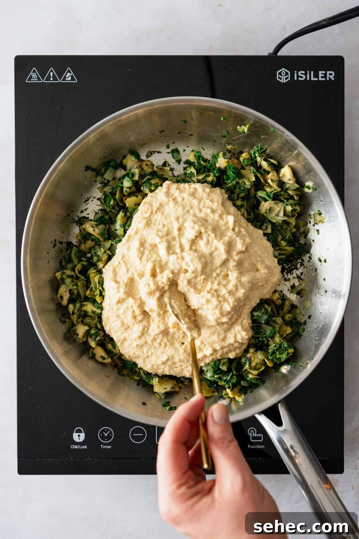 A pan with chopped artichoke hearts and spinach in it with a creamy sauce being spread over the vegetables.